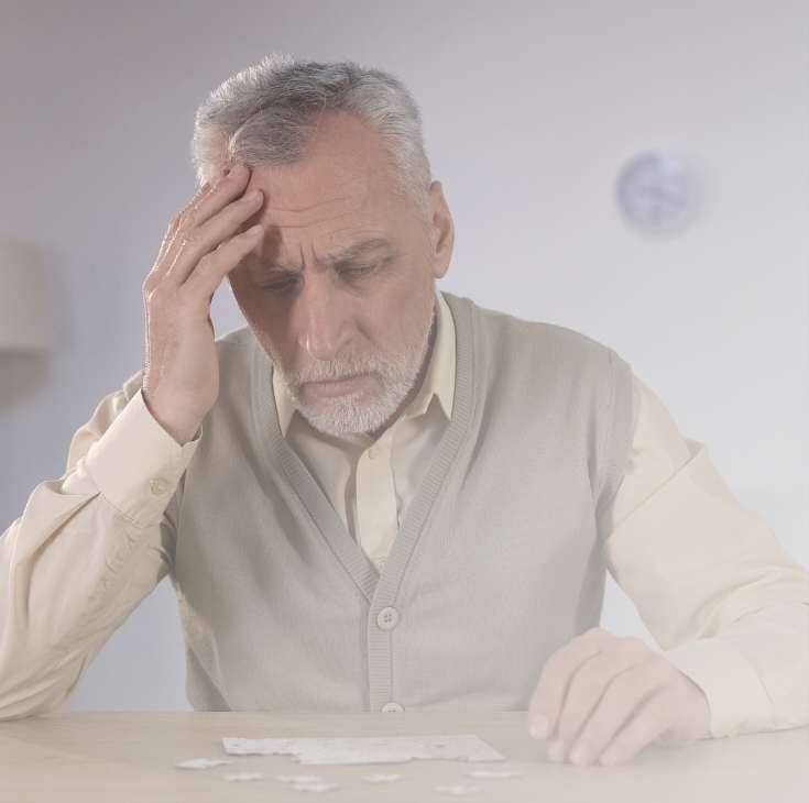 Starszy, siwy mężczyzna w beżowym kardiganie siedzi przy stole, trzymając się za czoło w geście głębokiego zamyślenia lub troski; przed nim na blacie leżą rozrzucone elementy białych puzzli. Zdjęcie jest utrzymane w jasnej, zamglonej tonacji.