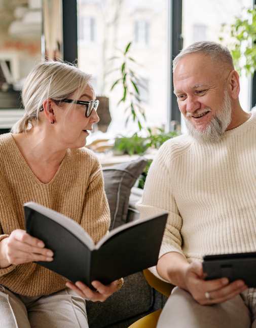 Uśmiechnięta para seniorów siedząca w jasnym salonie; kobieta w okularach trzyma otwartą czarną książkę lub notes i rozmawia z brodatym mężczyzną, który zadowolony spogląda na trzymany w dłoniach tablet.