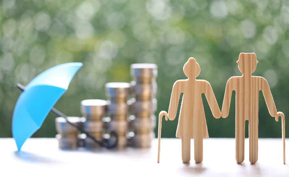 Drewniane figurki przedstawiające parę seniorów z laseczkami, stojące obok rosnących słupków z monet; nad monetami rozłożony jest mały, błękitny parasol ochronny. Całość znajduje się na rozmytym, zielonym tle natury.