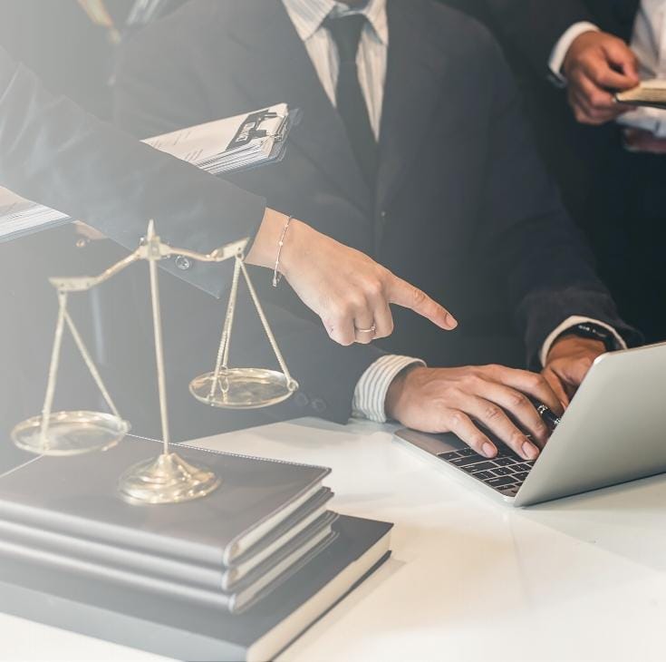 Zbliżenie na biurko w kancelarii, na którym stoją mosiężna waga szalkowa oraz stos grubych akt; w tle widać dłonie prawników pracujących przy laptopie – jedna osoba pisze na klawiaturze, a druga wskazuje palcem konkretny punkt na ekranie