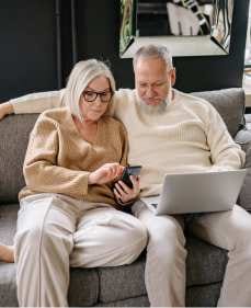 Starsza para siedzi blisko siebie na szarej kanapie; kobieta w okularach obsługuje smartfona, a mężczyzna obok niej trzyma na kolanach otwartego laptopa. Oboje ze skupieniem patrzą w ekrany urządzeń, wspólnie sprawdzając informacje.