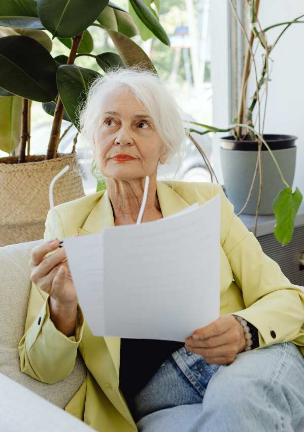 Starsza kobieta o siwych włosach, ubrana w jasnożółtą marynarkę, siedzi w otoczeniu roślin doniczkowych; trzyma w dłoniach dokumenty oraz okulary, spoglądając w górę z wyrazem głębokiego namysłu i determinacji.