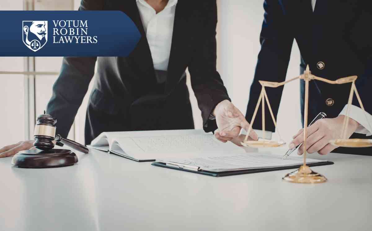 Two lawyers in suits review documents on a desk with a gavel and scales of justice; law firm logo in the corner.
